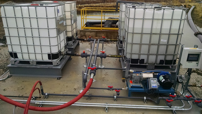 Four liquid polymer totes connected to a liquid polymer dosing skid setup next to a settling pond.
