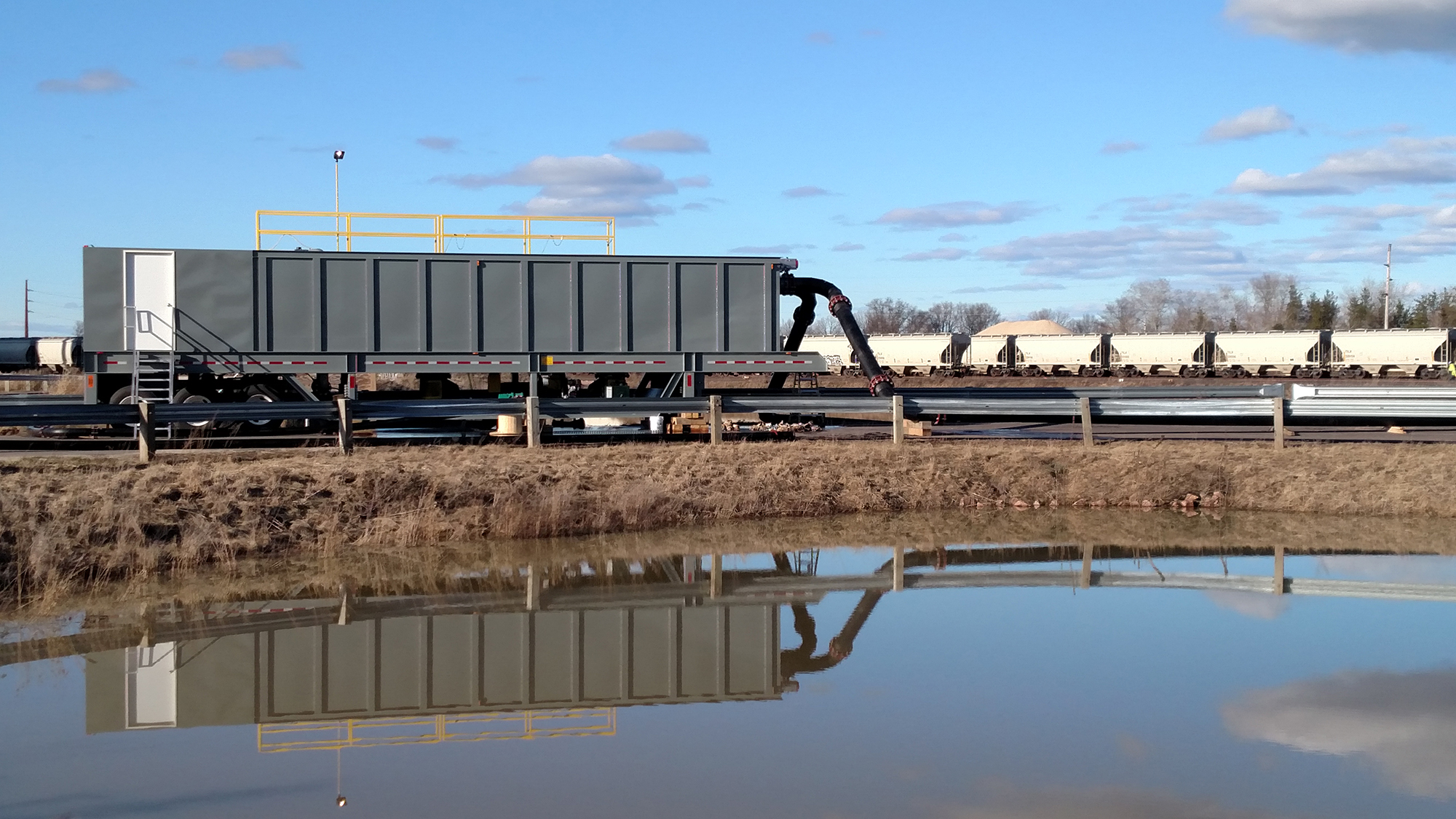 A grey rectangular portable water clarifier cribbed up next to a stormwater holding pond with a train in the background.