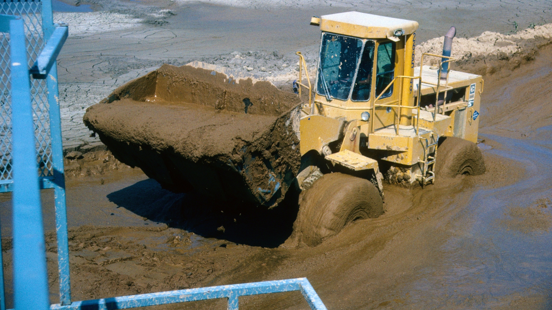 A front loader driving through a muddy settling pond with a watery bucket load.