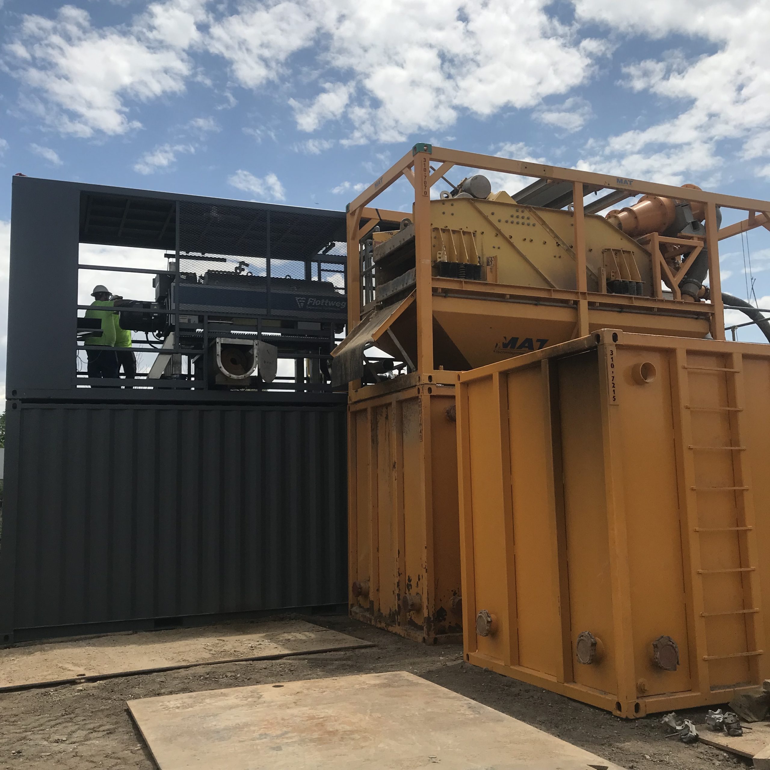 Large yellow containers next to a large grey container where two men stand next to a centrifuge on top of the grey container.