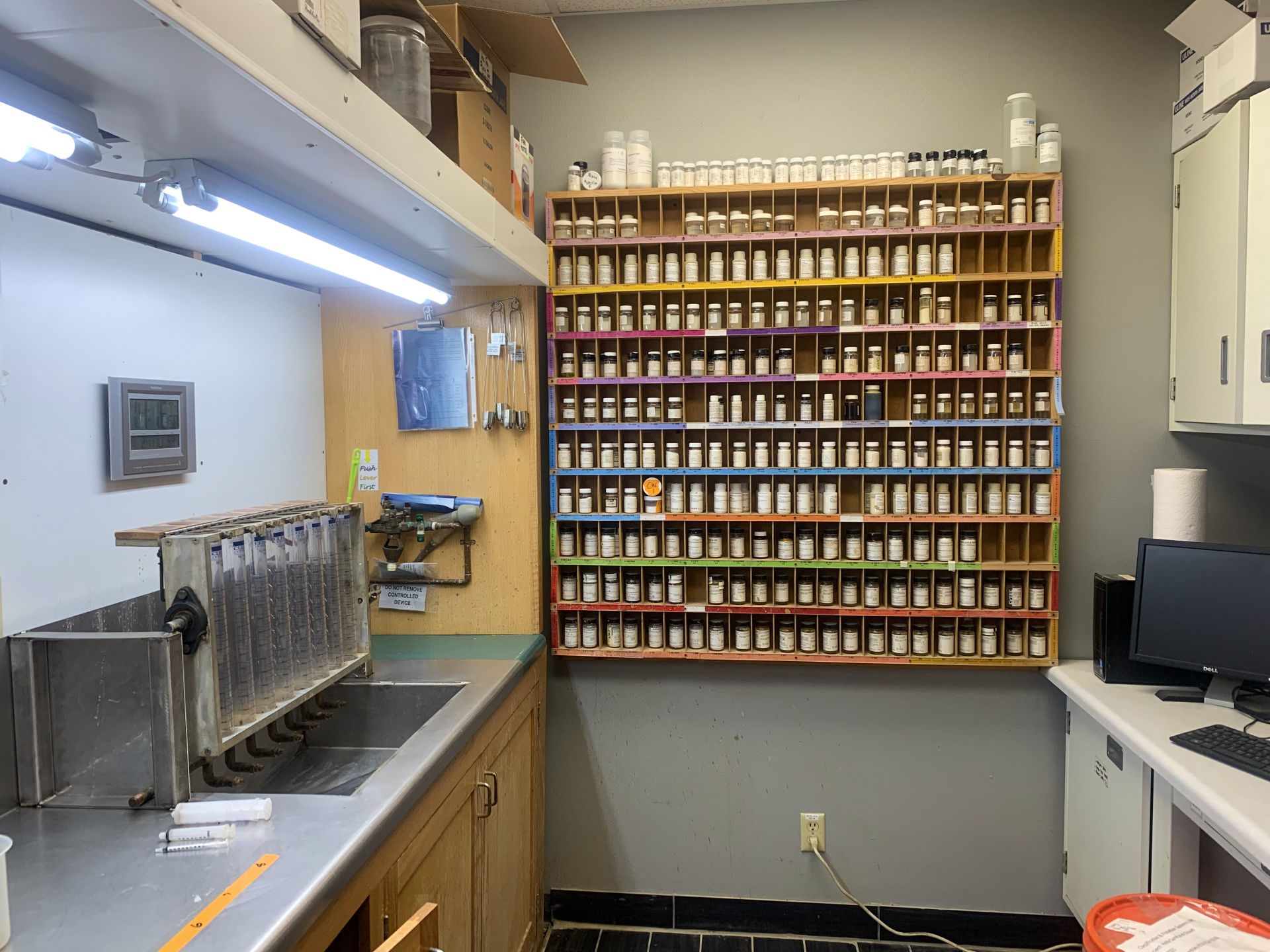 A small room with beakers over the sink of the left and a wall of different chemicals in the back.