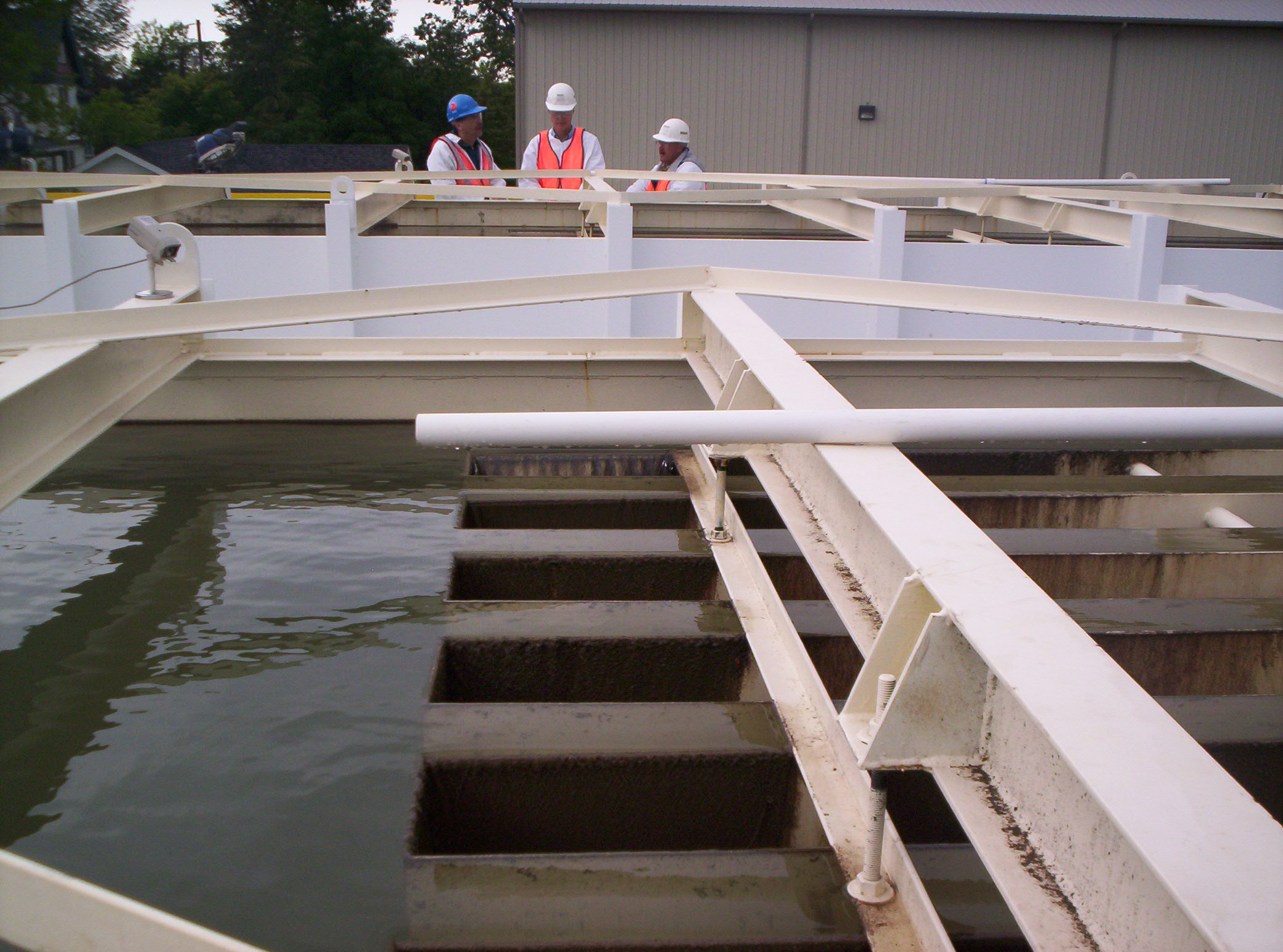 Three men in hard hats and safety vests looking at water overflow clarifier weirs.