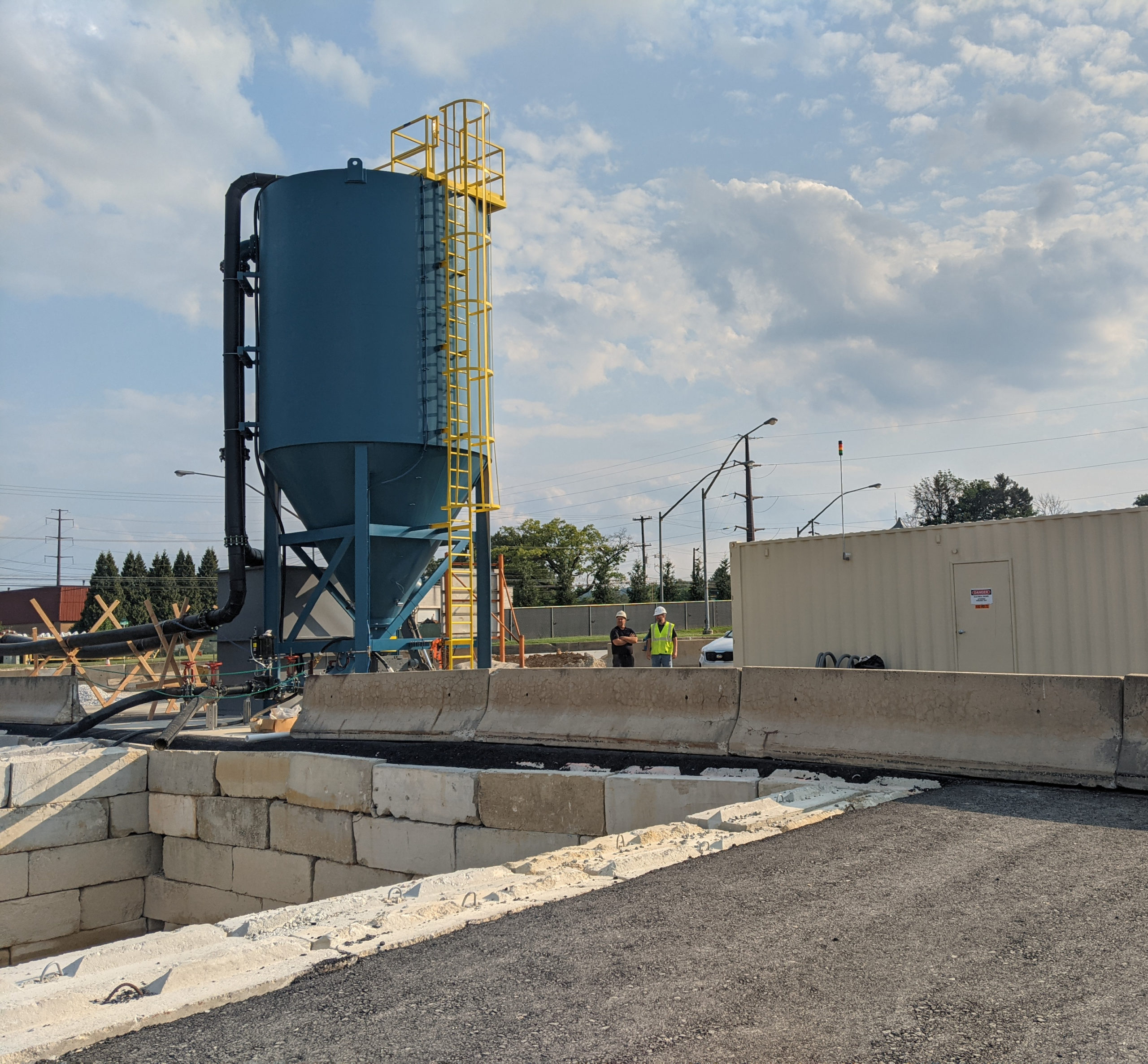 A vertical blue clarifier with workers to the right of it and a tan shipping container to the far right.