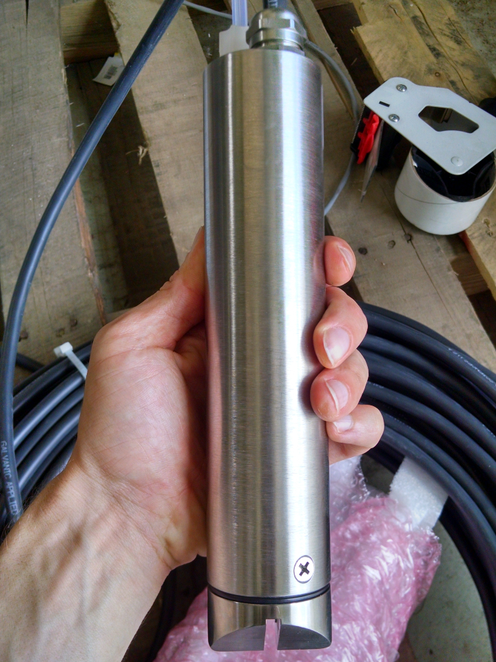 A stainless steel probe for water turbidity testing in someones hand with pallets and cords in the background.