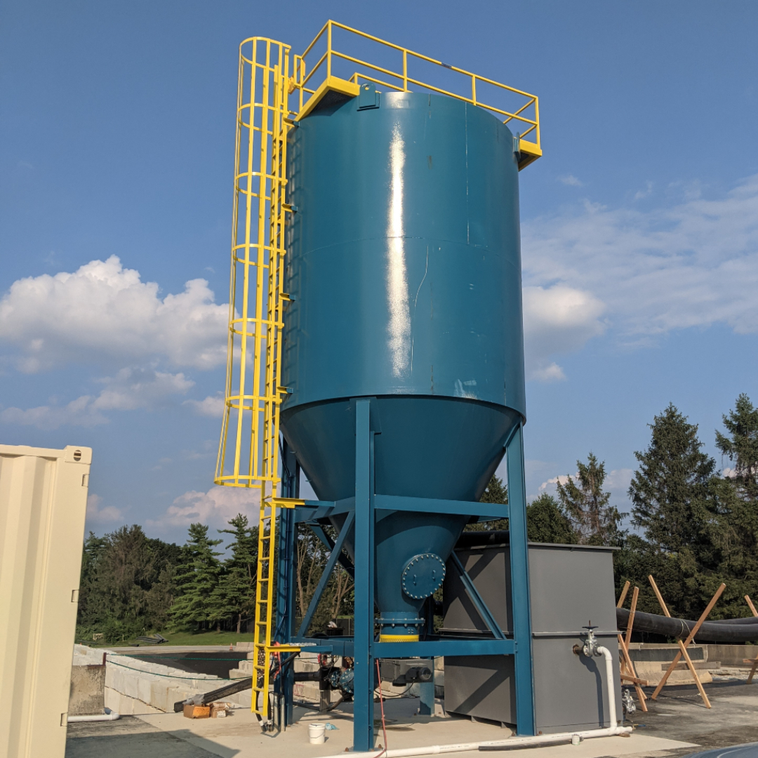 A large sun-soaked, blue, vertical, and cone bottom tank with a yellow fixed steel ladder and protection cage.