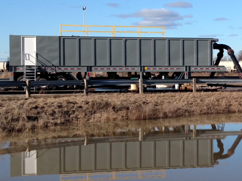 A grey water clarifier tank on wheels with a blue sky and stormwater pond in the foreground.