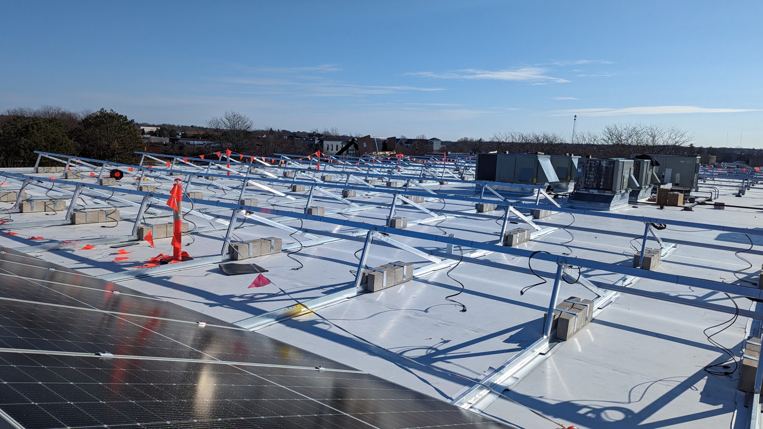 The top of an industrial building roof in the winter with solar panels, HVAC units, and construction flags in view.
