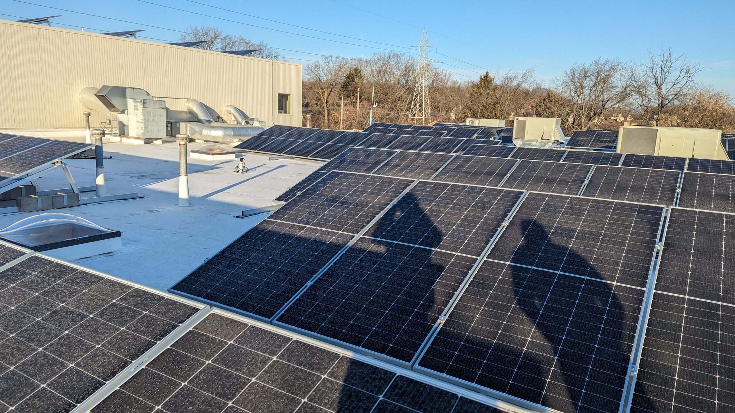 The face of solar panels on a roof with shadows across the panels and an industrial building side and trees in the background.