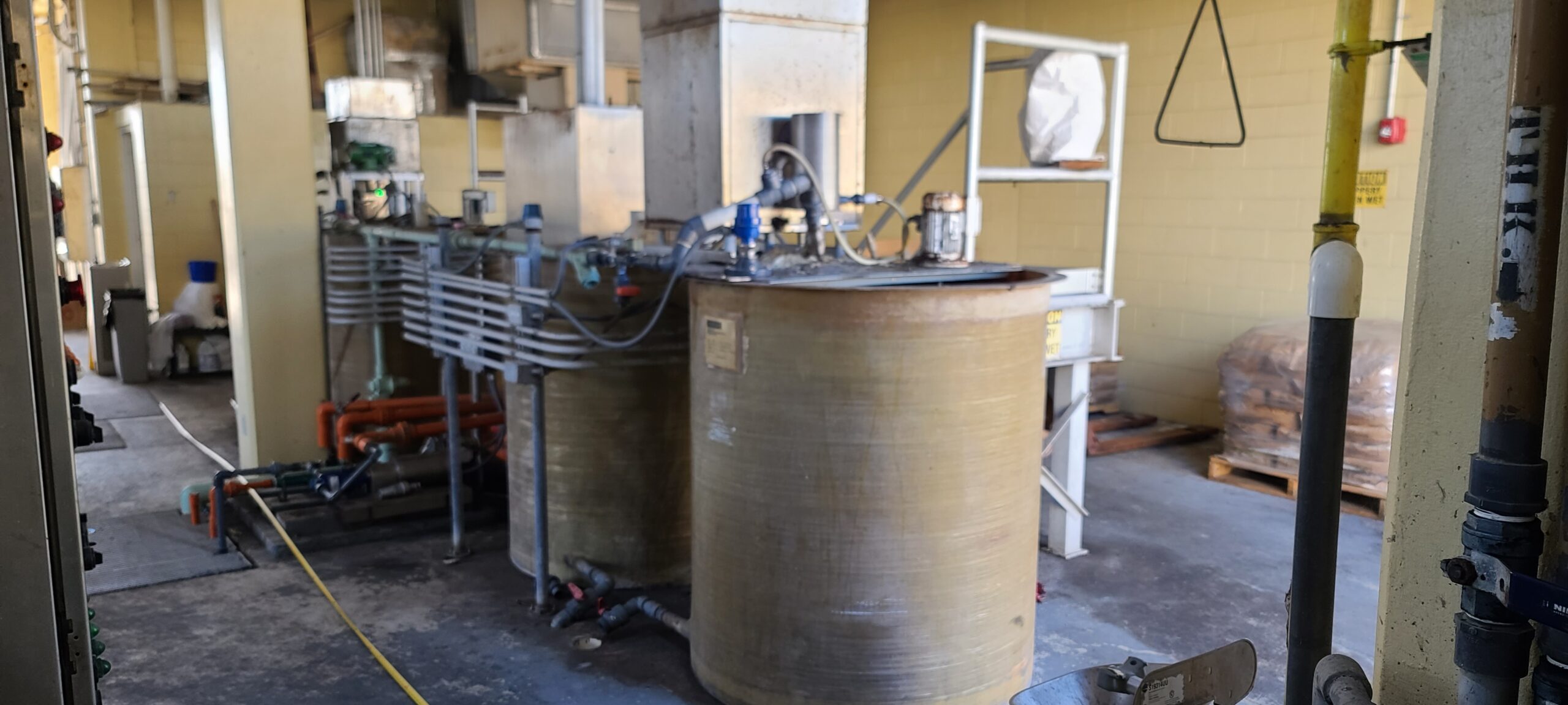 Customer Site 1 Two old round tanks with motorized impellers and outdated industrial steel equipment that create a run down polymer system. The equipment is inside a portion of a wastewater treatment building.