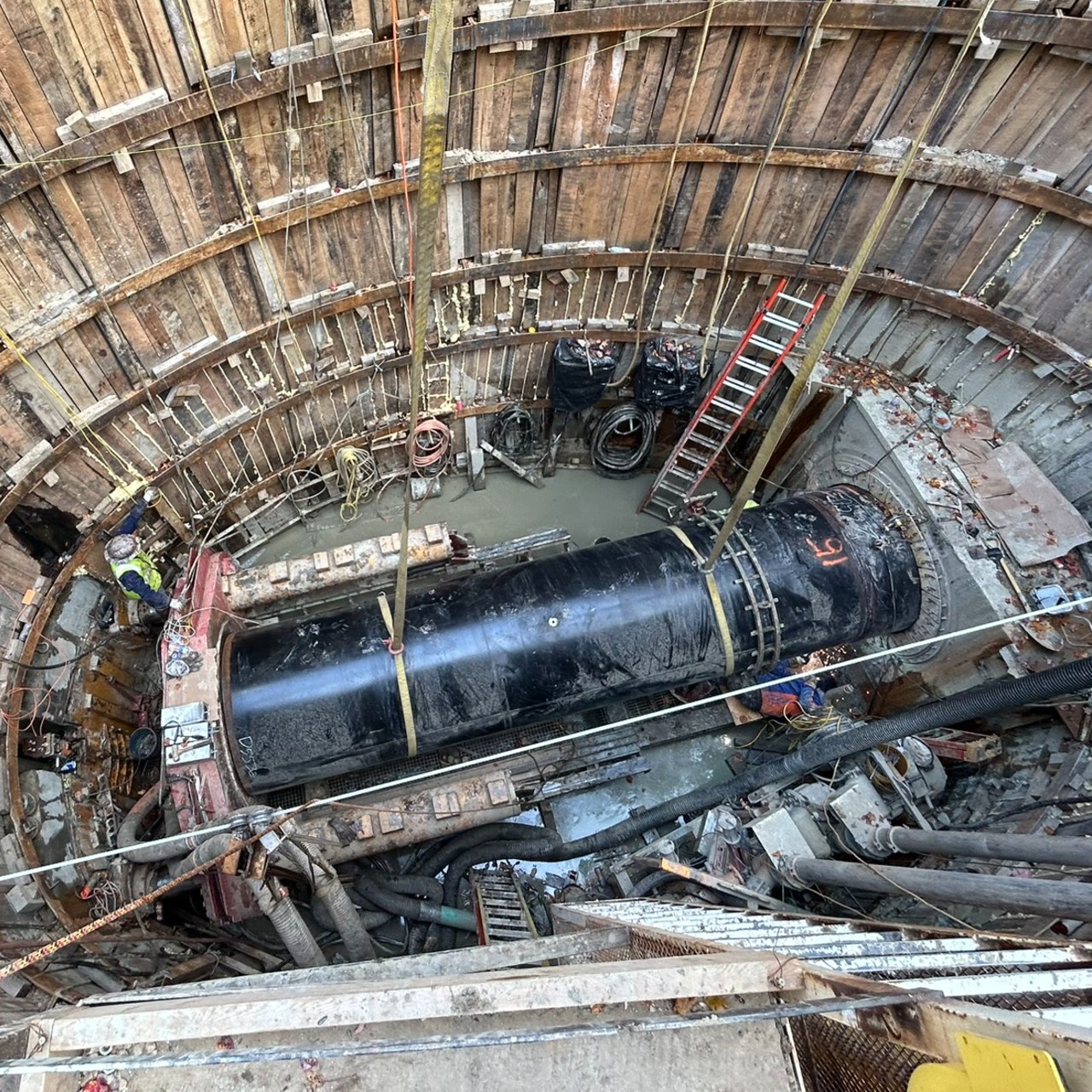 Tunnel – Microtunneling Looking down into a large hole at a construction site where there is a big black pipe supported by straps going into a tunnel.